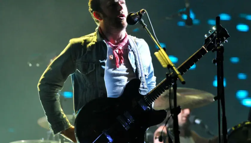 Wokalista Kings of Leon Polska na koncercie, gra na gitarze elektrycznej, w dżinsowej kurtce i czerwonej chuście.
