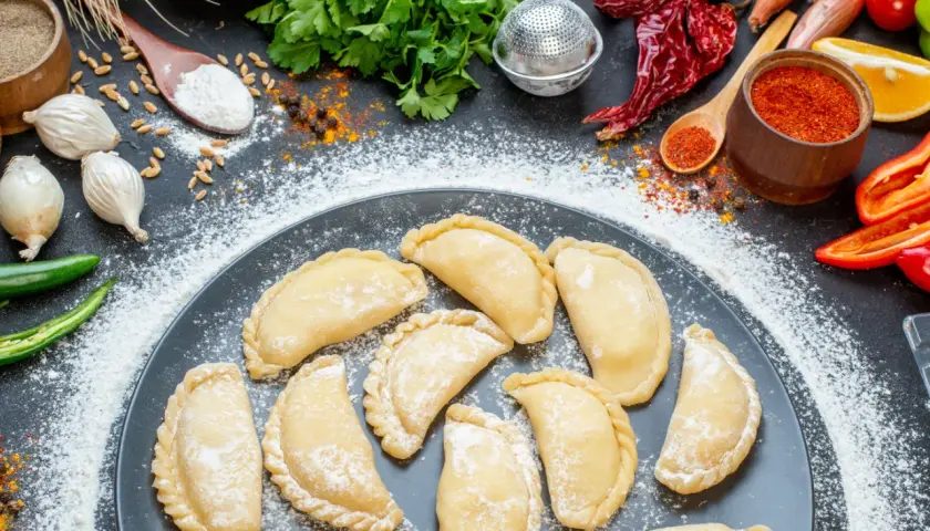 Gotowi, by nauczyć się, jak zrobić pierogi? Na talerzu surowe pierożki, obok mąka, cebula, papryczki chili i przyprawy.