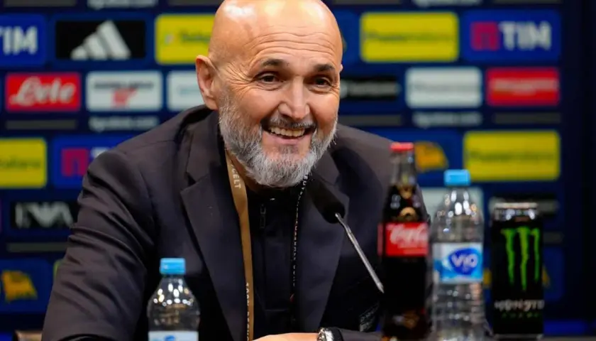 Uśmiechnięty trener Juventusu, z siwą brodą, siedzi przy stole z napojami.