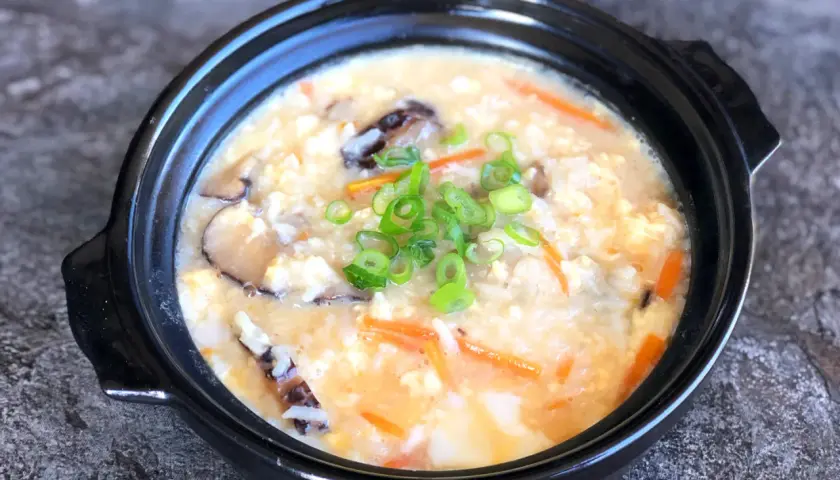 A comforting bowl of Japanese rice soup, also known as okayu, filled with tender rice, shiitake mushrooms, shredded carrots, and scrambled egg, garnished with fresh green onions.
