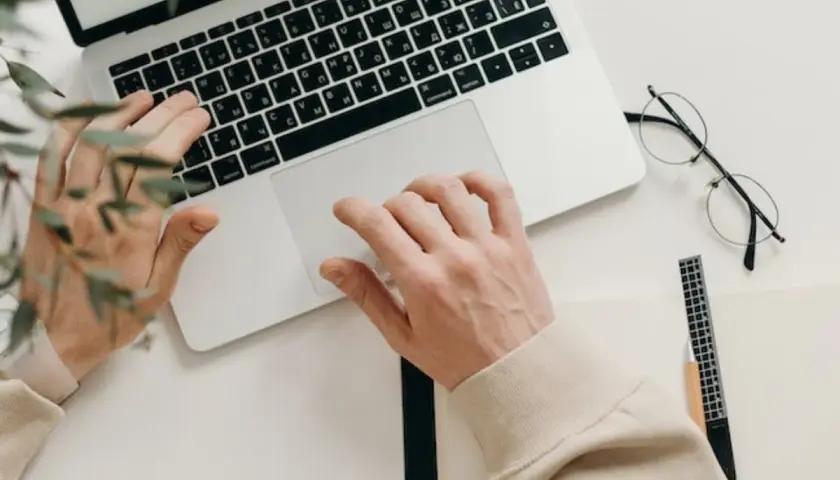 Ręce piszą na klawiaturze, obok leżą okulary i notatnik. Idealny tani laptop na studia, by zacząć naukę.