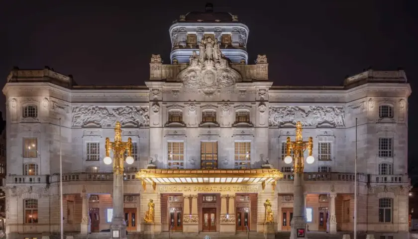 Kungliga Dramatiska Teatern i Stockholm, upplyst på natten. En storslagen byggnad med detaljerad fasad och gyllene utsmyckningar.