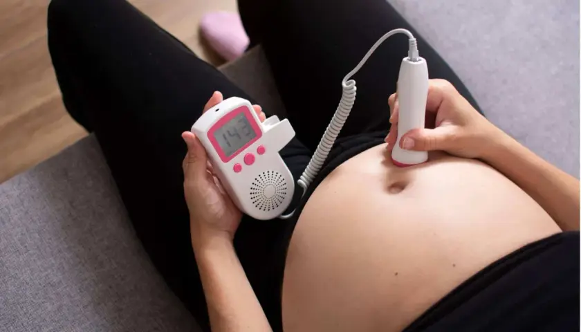 Una donna incinta usa un doppler fetale per ascoltare il battito del feto toccando la pancia. Il display mostra 143 bpm.