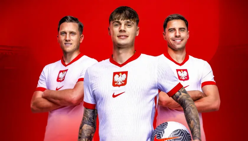 Trzech uśmiechniętych piłkarzy reprezentacji Polski w białych koszulkach z godłem orła. Jeden z nich trzyma piłkę.