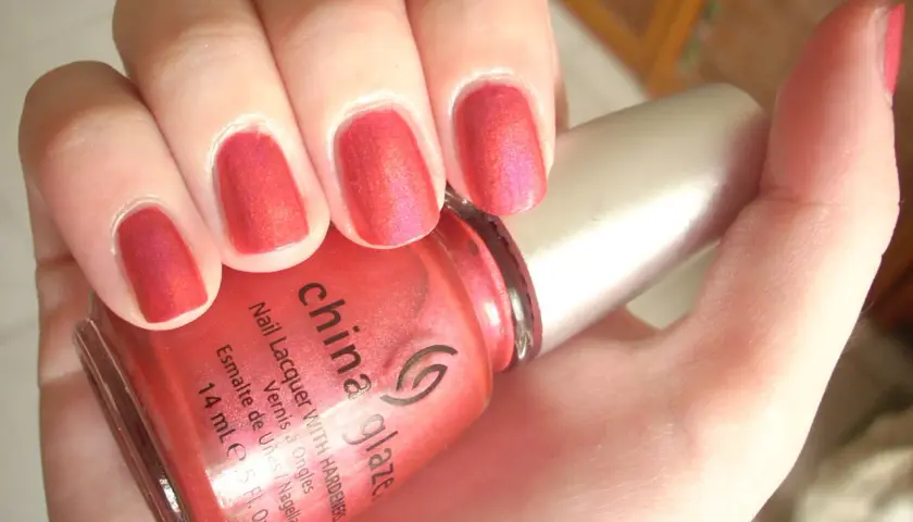 En hand med naglar målade i en glittrig, korallröd nyans. En flaska China Glaze nagellack syns i förgrunden.