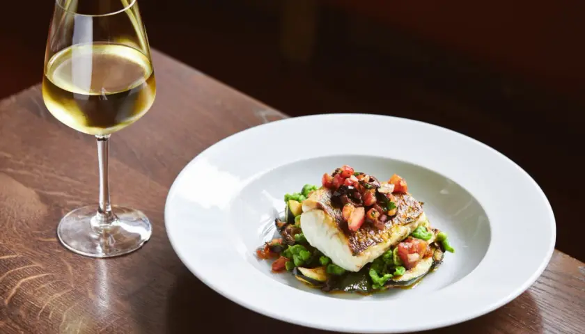 Un verre de vin blanc accompagne un poisson en sauce, servi sur un lit de légumes verts et de courgettes.
