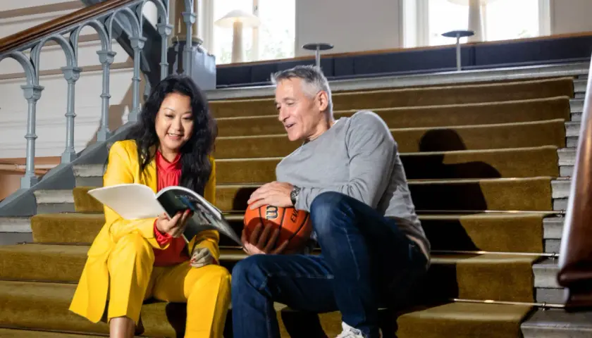 En kvinna i gul dräkt läser en bok medan en man med basket håller ett samtal. Kanske de diskuterar hur man kan jobba med att skriva om sport.