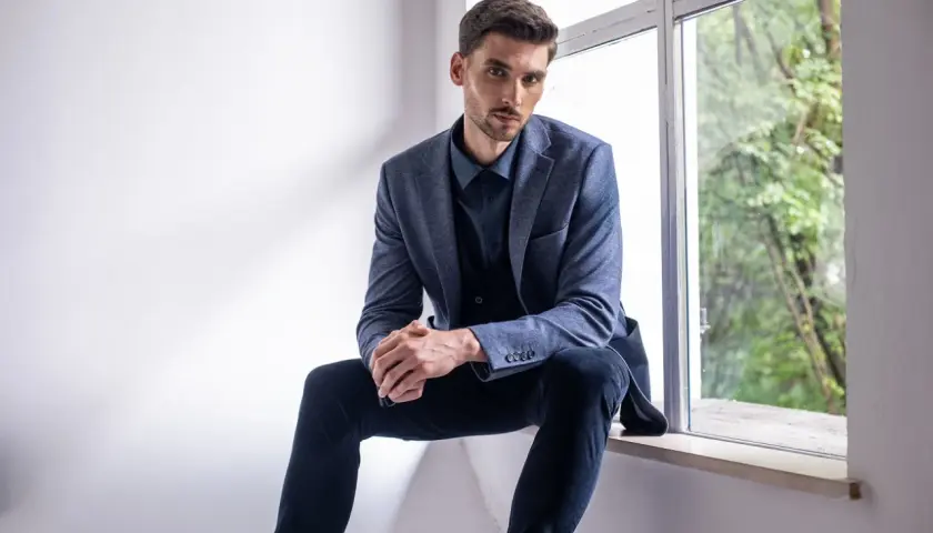 Mężczyzna w granatowym garniturze siedzi przy oknie. Jego elegancki strój podkreśla jego sylwetkę, a **kolor błękitny** marynarki dodaje mu klasy.