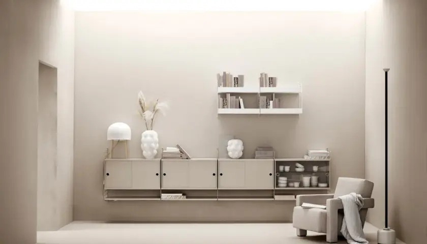Minimalistiskt vardagsrum med stringhylla, beigefärgad förvaring, fåtölj och lampa.