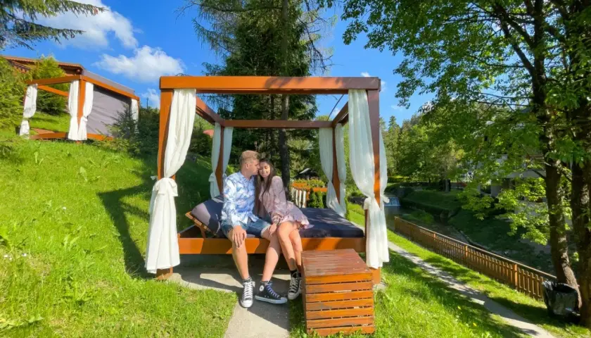 Idealny wypad na weekend we dwoje. Para siedzi przytulona na łóżku pod baldachimem, otoczona zielenią i strumykiem.