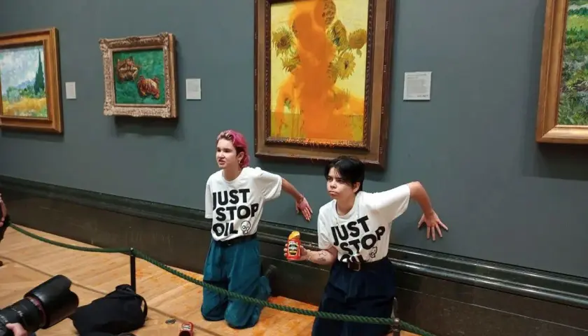 Dwie osoby w koszulkach "JUST STOP OIL" klęczą przed obrazem "Słoneczniki Van Gogha", na który wylano pomarańczową farbę.