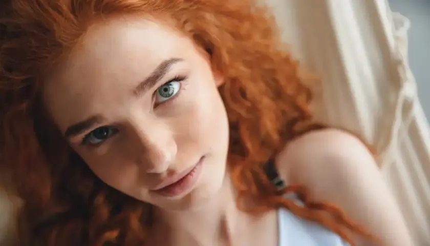 Primer plano de una joven con cabello pelirrojo natural y ojos claros, recostada en una hamaca.