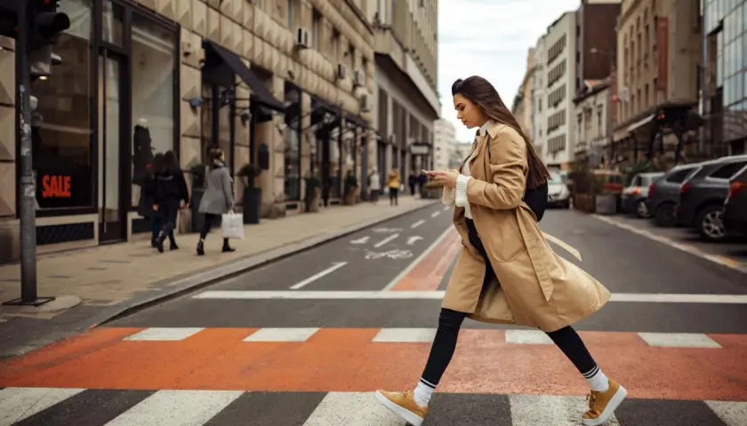 Kobieta w beżowym płaszczu i żółtych sneakersach przechodzi przez pasy. Idealne buty do trencza na miejski spacer.