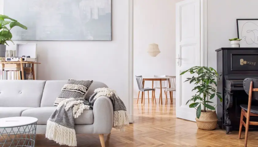 Un salón moderno con sofá gris, manta, cojín, mesa de café y piano negro. La luz blanca ilumina el espacio, creando una atmósfera acogedora.
