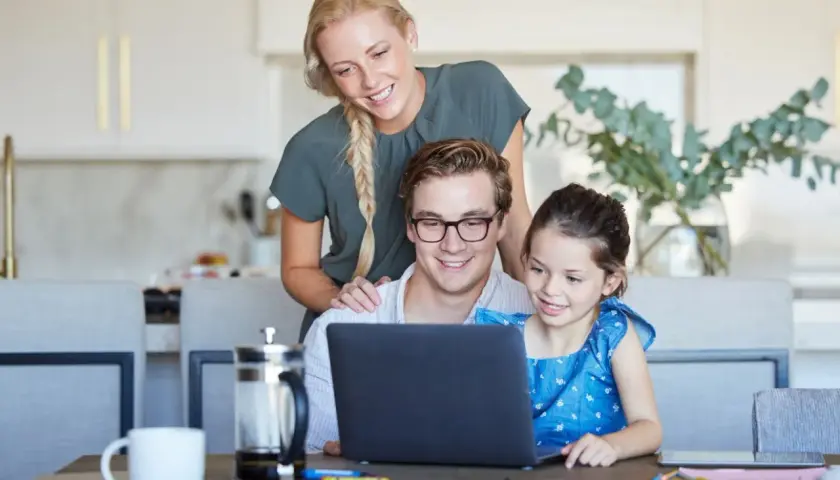 Rodzina cieszy się wspólnym czasem, tata na urlopie rodzicielskim uczy córkę korzystać z laptopa, mama wspiera ich z tyłu.