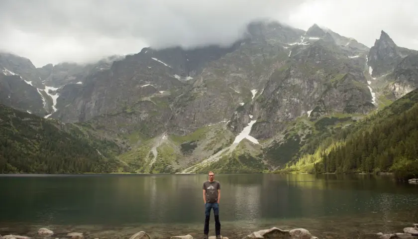 Mężczyzna stoi na kamieniach nad brzegiem jeziora Morskie Oko. W tle majestatyczne góry Tatr.