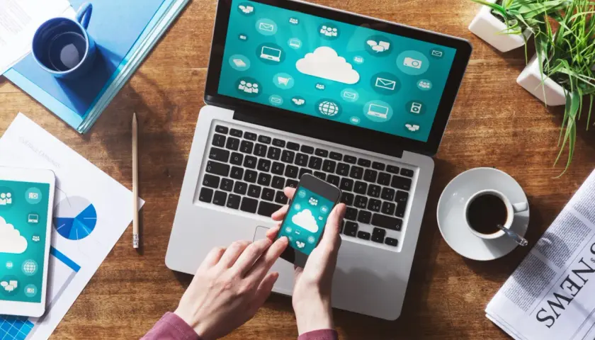 Ręce trzymają smartfon, który pokazuje ikony chmury. Obok laptop z podobnym ekranem. To inspiracja, jak połączyć komputer z internetem z telefonu.