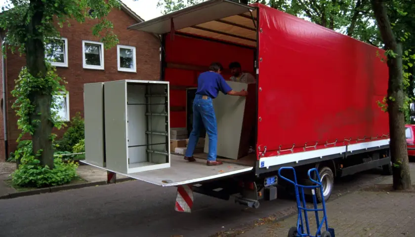 Umzugshelfer laden einen Schrank auf einen roten LKW. Der Schrank steht am Straßenrand, was die Bedeutung von "kerbside" verdeutlicht.
