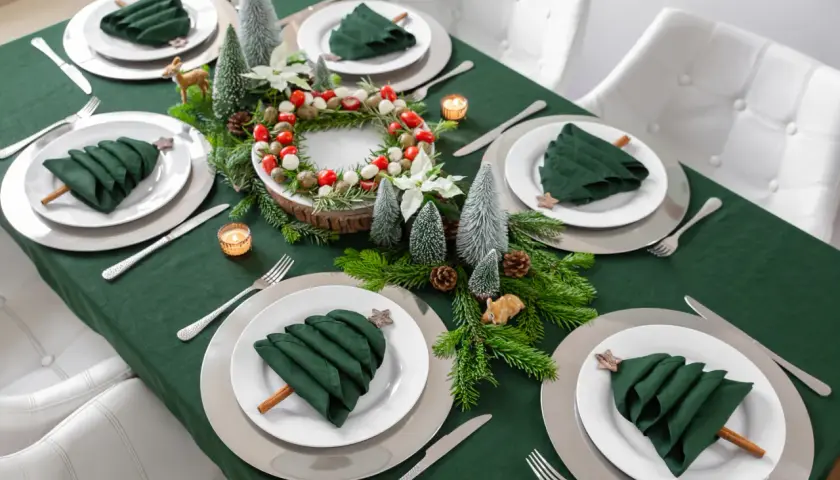 Moderne tischdeko weihnachten: grüne Servietten als Bäume, Tannenzweige, Kerzen und ein Kranz mit Beeren auf einem grünen Tischtuch.