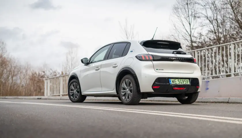 Biały Peugeot e-208 z zieloną tablicą rejestracyjną WE 5G959 zaparkowany na drodze.