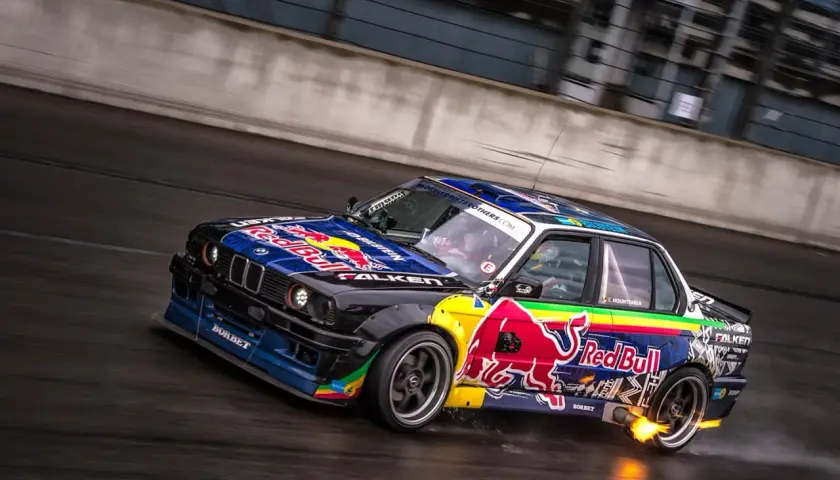 BMW E30 w barwach Red Bull, auto do driftu, z płomieniami z wydechu, na mokrym torze.
