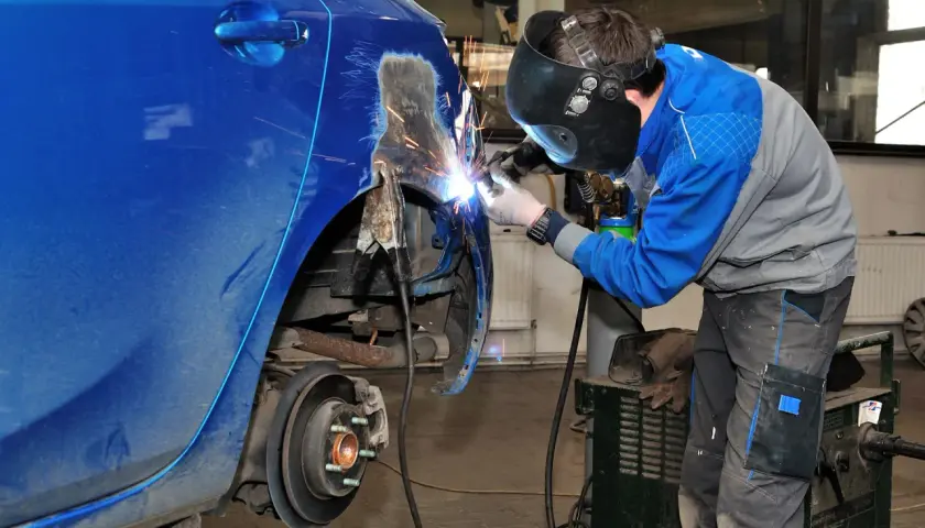 Mechanik w masce spawa blachę samochodową, przywracając jej dawny kształt. Iskry sypią się podczas prac blacharskich.