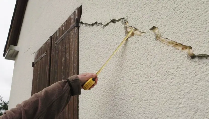 Une main mesure une large fissure sur le mur extérieur d'une maison, près de volets en bois.