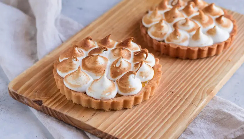 Due crostatine con crema al limone e meringa all'italiana, dorata e soffice, su un tagliere di legno.