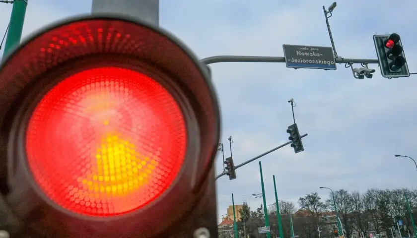 Zbliżenie na czerwone światło sygnalizatora, obok widać rejestrator przejazdu na czerwonym świetle i znak ronda Jana Nowaka-Jeziorańskiego.
