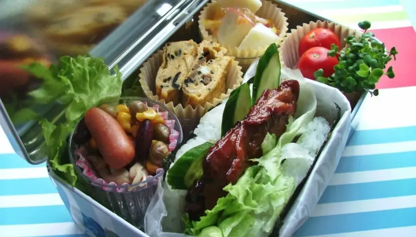 A delicious bento box with a glazed meat skewer, rice, lettuce, egg roll, sausage, beans, corn, tomatoes, and sprouts.