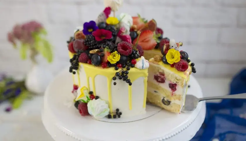 Wyjątkowy jesienny tort udekorowany świeżymi owocami, kwiatami i bezami. Kawałek ciasta już czeka na widelcu.