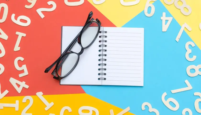 Occhiali neri su un quaderno aperto, circondati da numeri in legno sparsi su sfondi colorati. Un promemoria visivo per imparare l'inglese, magari il numero 66.