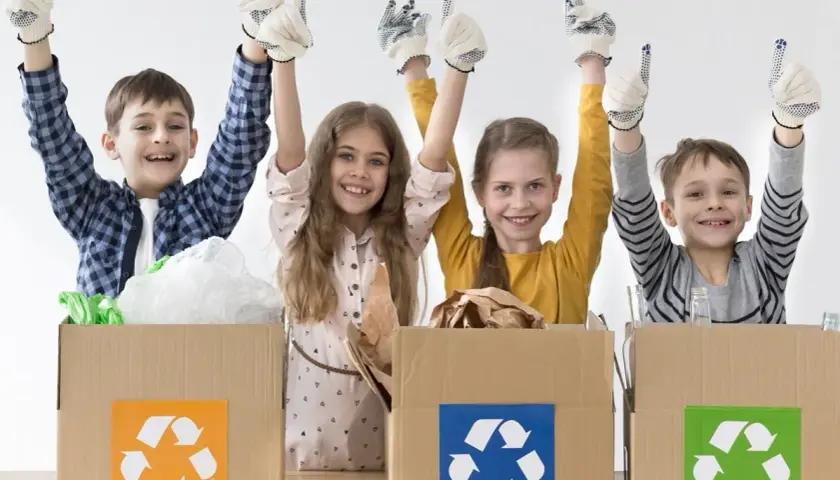 Niños felices participan en un taller de reciclaje, con guantes y pulgares arriba, junto a cajas de cartón con símbolos de reciclaje.