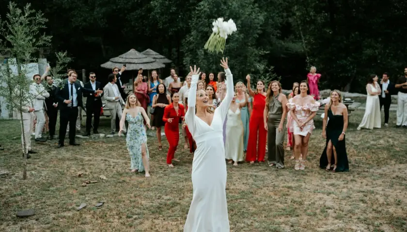 Panna młoda w białej sukni rzuca bukietem, a zgromadzeni goście, w tym panie w kolorowych kreacjach, czekają na jego złapanie. To piękny moment celebrujący tradycje weselne.