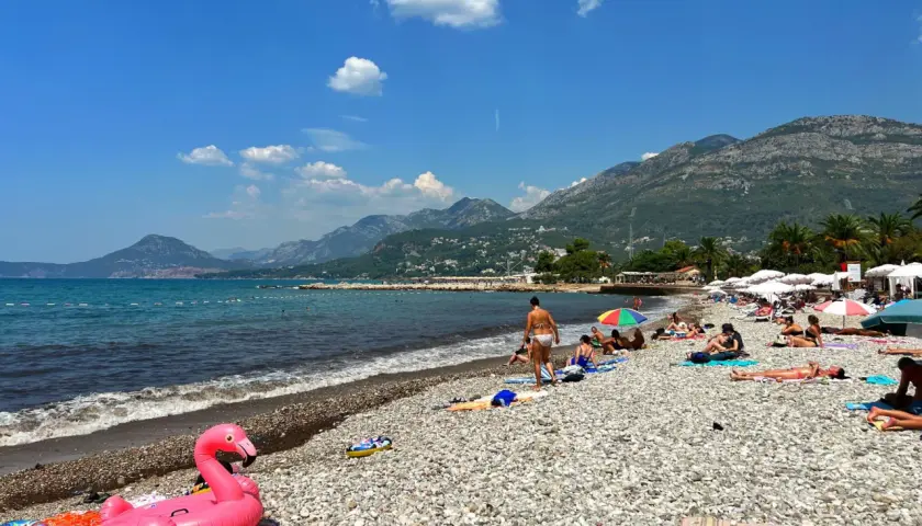 Słoneczny dzień na plaży w Barze, Czarnogóra. Ludzie odpoczywają na kamienistej plaży, a w tle widać góry. Różowy flaming unosi się na wodzie.