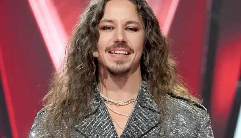 Michał Szpak w błyszczącej kurtce i z długimi włosami, uśmiecha się do kamery.