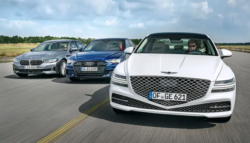 Un Genesis blanco lidera un grupo de sedanes de lujo, incluyendo un Audi azul y un BMW gris, en una carretera abierta.