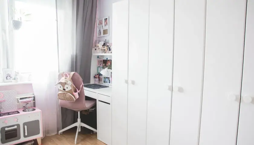Urocza aranżacja małego pokoju z dużą białą szafą, biurkiem, różową kuchnią dla dzieci i plecakiem w kształcie sowy.