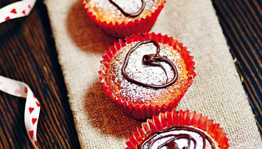 Tre saffransmuffins med mandelmassa, pudrade med florsocker och dekorerade med choklad. En vit sidenband med hjärtan ligger bredvid.