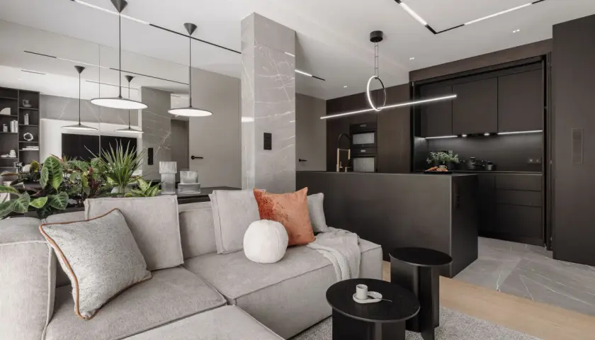 Nowoczesny salon z kuchnią, gdzie oświetlenie wpuszczane tworzy elegancką atmosferę. Szara sofa, czarne meble kuchenne i marmurowe detale.