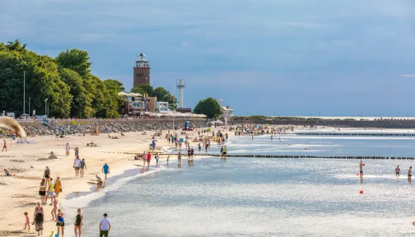 Tłumy ludzi na piaszczystej plaży, latarnia morska w tle. Idealne miejsce na tanie miejscowości nad morzem.