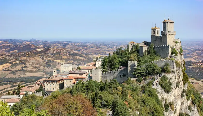 Widok na twierdze i miasto San Marino na wzgórzu, z pagórkowatym krajobrazem w tle.