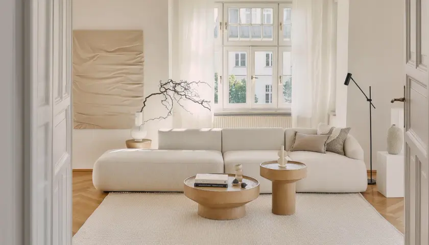 Jasny, przytulny salon z beżowym domem w tle. Miękka sofa, drewniane stoliki i subtelne dekoracje tworzą spokojną atmosferę.
