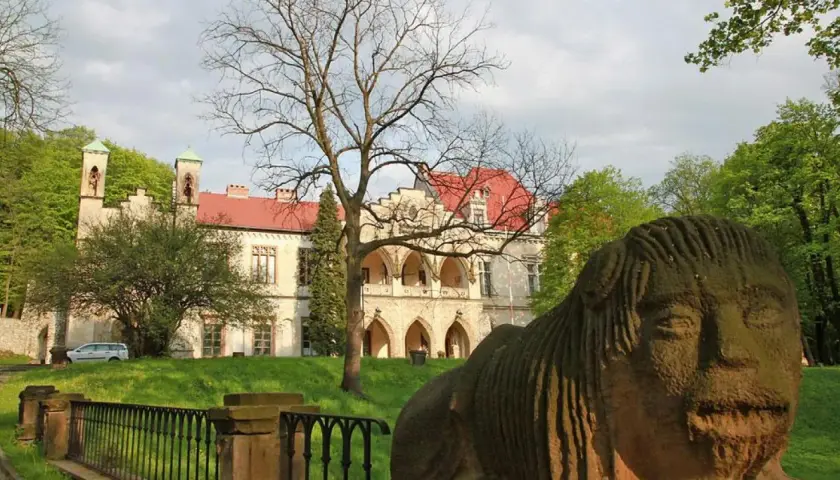 Kamienna rzeźba twarzy przed pałacem w Młoszowej. Budynek z czerwonym dachem i arkadowym gankiem otoczony zielenią.