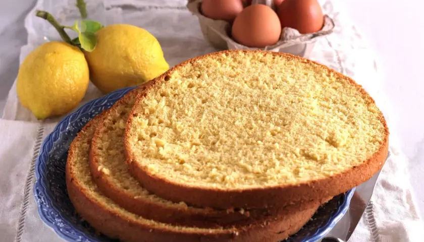 Fette di soffice sponge cake su un piatto blu, con limoni e uova sullo sfondo, pronte per una deliziosa torta.