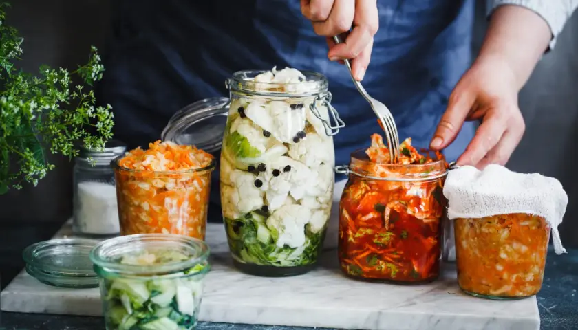 Fermentiertes Gemüse in Gläsern: Blumenkohl mit Pfefferkörnern, Karotten-Sauerkraut und ein Glas mit rotem Kimchi, das mit einer Gabel bearbeitet wird.