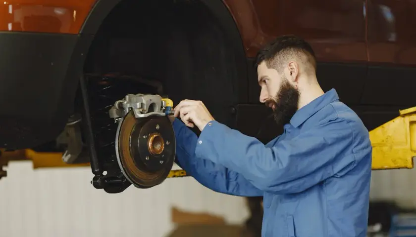 Mechanik w niebieskim kombinezonie pracuje przy hamulcach samochodu. Widać tarczę hamulcową i zacisk. To ważny punkt, od dot 3 a dot 4 zależy bezpieczeństwo jazdy.
