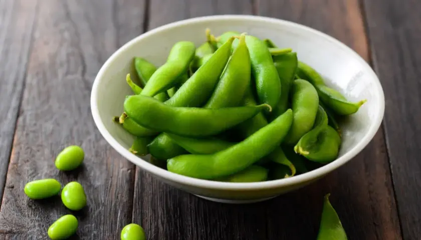 Miseczka pełna zielonej fasolki edamame na drewnianym tle. Kilka fasolek rozsypanych obok.