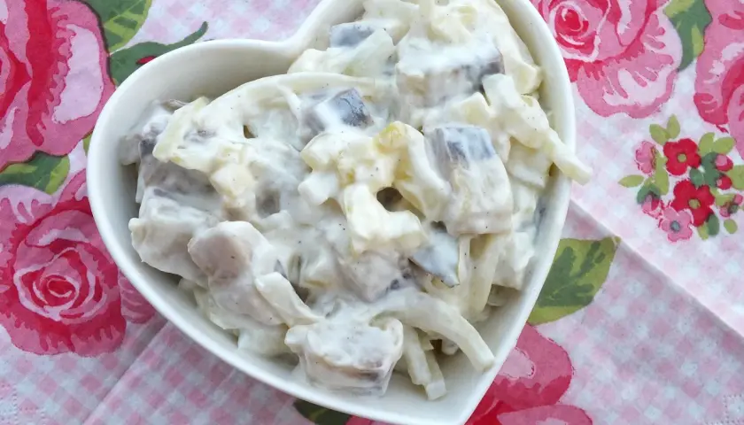 Pyszny przepis na śledzie w śmietanie z jabłkiem i ogórkiem. Sałatka w białej miseczce w kształcie serca na tle różowych róż.