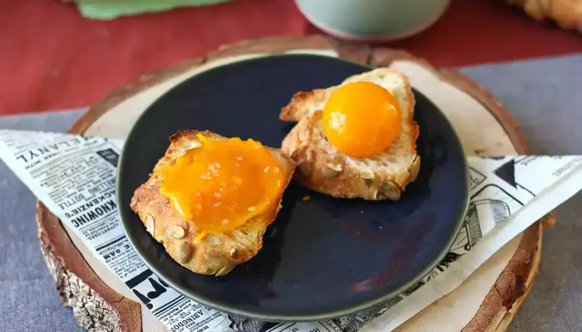 Sur une assiette, deux toasts de pain aux graines, l'un garni d'une texture orangée et l'autre d'un jaune d'œuf confit brillant.